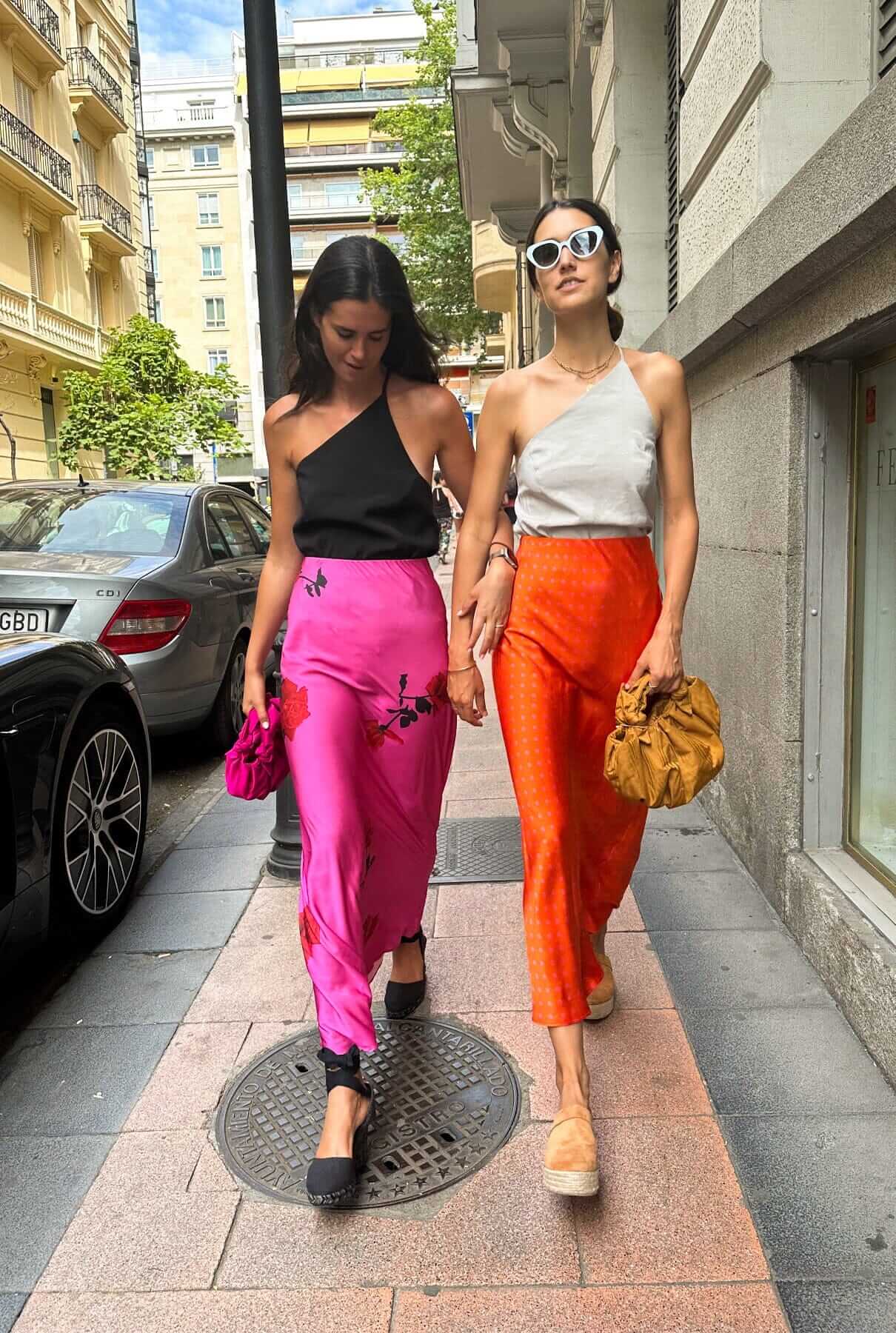 Two women wearing stylish skirts, one in pink floral and the other in orange, showcasing trendy skirts on the street.